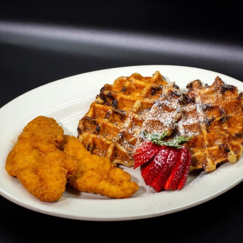 Golden chicken and waffles with powdered sugar and sliced strawberries on a white plate.